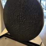 Bronzeskulptur in Form einer Scheibe mit dunkler, oxidierter Oberfläche, aufgenommen von schräg hinten auf einem hellen Holztisch. Die Struktur zeigt viele kleine, unregelmäßige Vertiefungen. Im Hintergrund ist ein Galerieraum sichtbar.