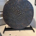 Bronzeskulptur „Scheibe“ von Herbert Baumann frontal aufgenommen. Die dunkel patinierte Oberfläche ist von zahlreichen Erhebungen und Vertiefungen durchzogen, mittig verdichtet. Die Skulptur steht auf einem schwarzen Metallständer vor heller Wand.